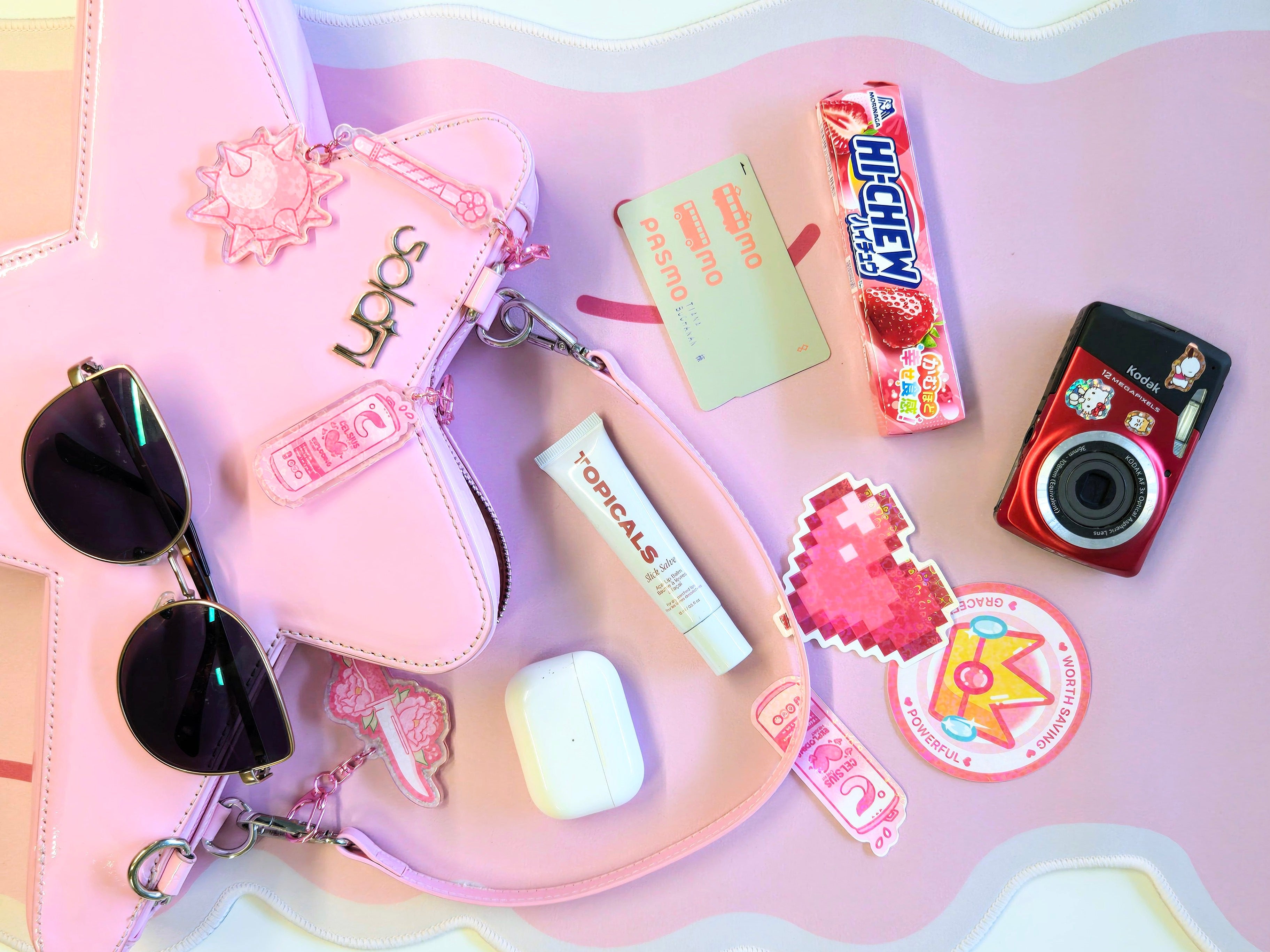 Collection of small items including a camera, candy, lip balm, and sunglasses on a pink surface with decorative elements.