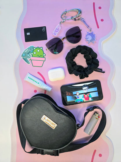 Collection of small items including a black bag, phone, sunglasses, and scrunchie on a pink surface with cartoon designs.